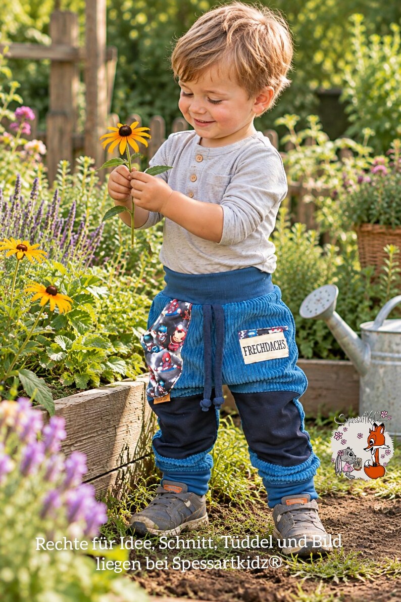 May include: A light gray long-sleeve henley shirt paired with blue corduroy pants featuring a superhero print pocket, a patch labeled FRECHDACHS, dark blue knee panels, and ribbed cuffs. The pants have a drawstring waist and a relaxed fit.