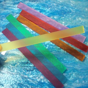 May include: A set of six frosted glass nail files in various colors, including purple, red, orange, yellow, green, and blue. The files are arranged in a rainbow pattern and are lying on a blue surface with glitter.