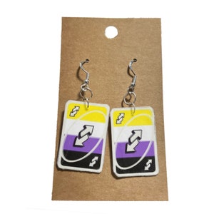 May include: A pair of handmade earrings featuring a reverse card design with yellow, white, purple, and black colors. The earrings are attached to silver-colored fish hook ear wires. The earrings are displayed on a brown card.