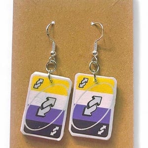 May include: A pair of rectangular dangle earrings featuring a graphic design of a game card with yellow, white, purple, and black horizontal stripes and a central white arrow icon. The earrings are attached to silver-toned metal hooks.