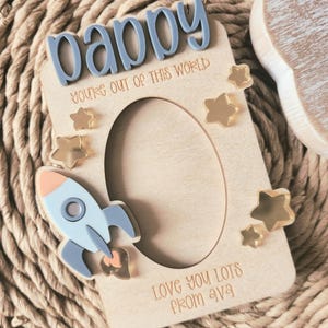 May include: Wooden photo frame with the word "Daddy" in blue letters. The frame has a rocket ship and star accents. Text on the frame reads "You're out of this world" and "Love you lots from Ava".