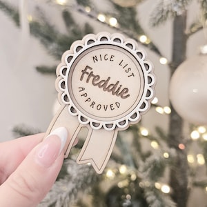 May include: A wooden Christmas decoration shaped like a badge, with a scalloped edge and two ribbons. The text "NICE LIST Freddie APPROVED" is engraved on the front. The ornament is held in front of a Christmas tree with fairy lights.