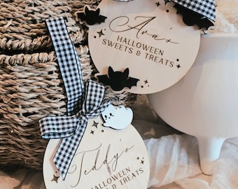 Puede incluir: Dos adornos redondos de madera de Halloween con lazos de cuadros. Uno dice "Ava's Halloween Sweets & Treats" y presenta murciélagos y estrellas recortadas. El otro dice "Teddy's Halloween Sweets & Treats" con fantasmas y estrellas recortadas.