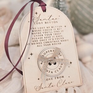 May include: Wooden Christmas ornament with a claret ribbon. The ornament reads "Santa's Lost Button" and features a silver bow and button. The text on the ornament asks the finder to keep it safe.
