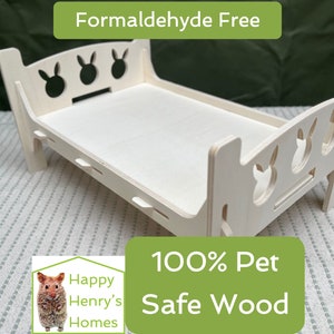 May include: A white wooden bed for small pets, featuring bunny ear cutouts on the headboard and footboard. The bed is raised off the ground on four legs. The text "Formaldehyde Free" and "100% Pet Safe Wood" is visible in the image.
