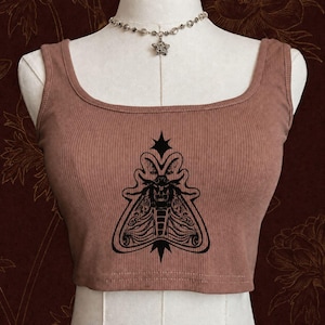 Può includere: Un top corto a coste color rosa polvere con una stampa grafica nera di una falena centrata sul petto. Il top presenta uno scollo rotondo e spalline larghe. Una collana a catena color argento con un ciondolo a forma di stella poggia sullo scollo.