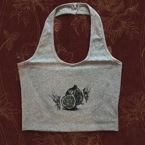 May include: A heather gray halter top featuring a black ink-style graphic of a sliced fig and two whole figs centered on the front. The garment has a wide, rounded neckline and a sleeveless design, constructed from a soft, jersey-knit fabric.