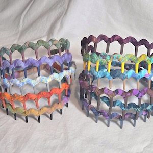 May include: Two decorative headbands with a layered design. Each headband features a series of colorful fabric loops attached to a frame. One headband has orange, yellow, and blue tones, while the other has purple and green tones.