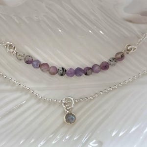 Op de afbeelding: Een gelaagde kettingenset bestaande uit een zilveren ketting met een rij kleine, gefacetteerde, paarse en gespikkelde kralen, en een tweede zilveren bolletjesketting met een kleine, ronde, grijsblauwe stenen hanger.