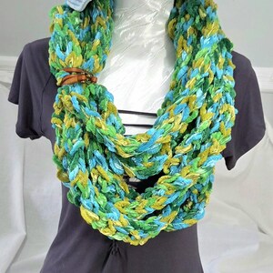 May include: A chunky infinity scarf featuring a blend of bright green, lime, and turquoise yarn loops. The scarf is secured with a small brown leather strap and a metallic button. A light blue denim bucket hat sits above the scarf on a dark shirt.
