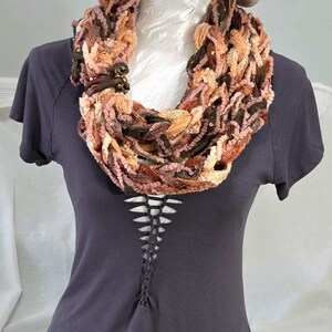 May include: A beige and white checkered bucket hat, a multi-textured infinity scarf in shades of pink, brown, and orange with a small metal clasp, and a dark gray short-sleeved t-shirt featuring a vertical lace-up detail at the center chest.