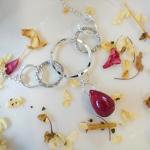 Puede incluir: Un collar de plata con múltiples círculos entrelazados y un colgante rojo en forma de lágrima.