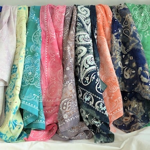 May include: A collection of square fabric bandanas arranged in a row, featuring a variety of colors including red, blue, yellow, teal, pink, grey, orange, green, and brown, all decorated with traditional white paisley and floral patterns.