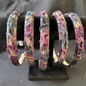 Puede incluir: Cinco diademas de tela sobre un expositor negro. Las diademas están envueltas en un material texturizado con una mezcla de colores que incluyen morado, azul, dorado y beige. Las diademas se muestran sobre un fondo negro.