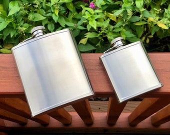 Peut inclure: Deux flasques en acier inoxydable, l'une plus grande que l'autre, sont présentées sur une surface en bois. Les flasques sont toutes deux brillantes et ont un design simple.