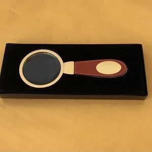 May include: A magnifying glass with a gold-coloured frame and a burgundy leather handle, presented in a black box. The handle has a gold-coloured oval plate. The magnifying glass is designed for reading or detailed work.