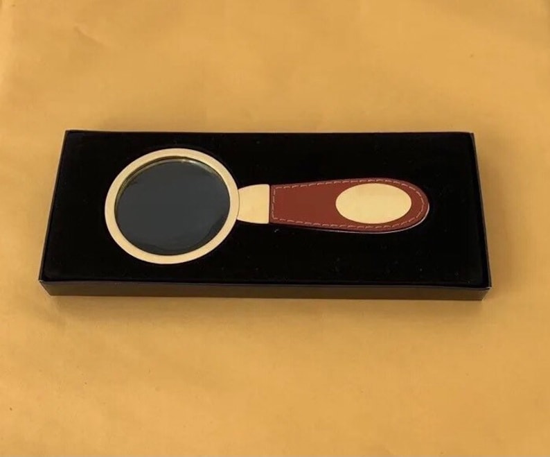 May include: A magnifying glass with a gold-coloured frame and a burgundy leather handle, presented in a black box. The handle has a gold-coloured oval plate. The magnifying glass is designed for reading or detailed work.