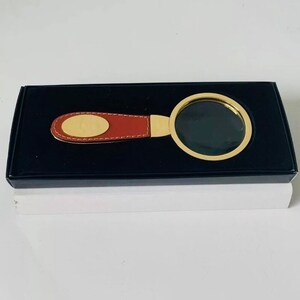 May include: A magnifying glass with a gold-coloured frame and a brown leather handle, presented in a black box. The handle features a gold-coloured oval accent. The magnifying glass is designed for reading and detailed viewing.