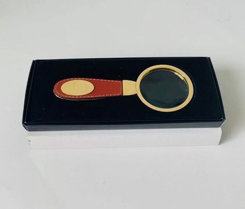 May include: A magnifying glass with a gold-coloured frame and a brown leather handle, presented in a black box. The handle features a gold-coloured oval accent. The magnifying glass is designed for reading and detailed viewing.