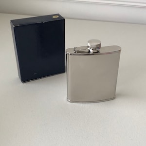 May include: A silver stainless steel hip flask with a screw-on cap. The flask is in a blue box.
