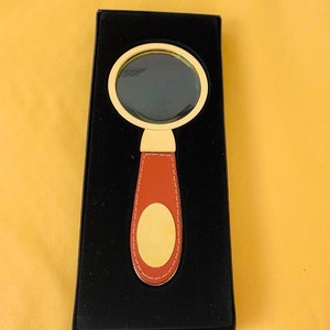 May include: A magnifying glass with a gold frame and a brown leather handle. The handle features a gold oval accent. The magnifying glass is presented in a black box against a yellow background.