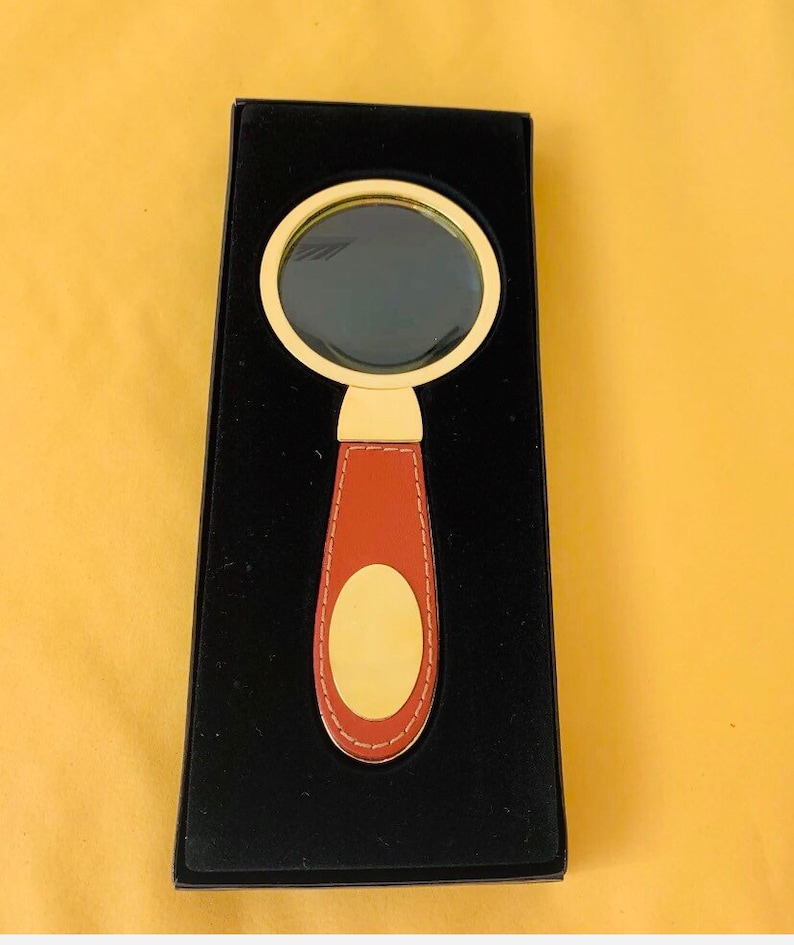 May include: A magnifying glass with a gold frame and a brown leather handle. The handle features a gold oval accent. The magnifying glass is presented in a black box against a yellow background.