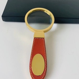 May include: A gold-coloured magnifying glass with a clear lens and a brown leather handle. The handle has a gold-coloured oval accent and is stitched with white thread. A black box is in the background.