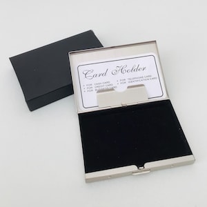 May include: A silver-toned metal card holder with a black interior and a black box. The card holder is open, displaying a card with the words "Card Holder" and various card types. The holder is designed to hold cash, credit, business, telephone, and identification cards.