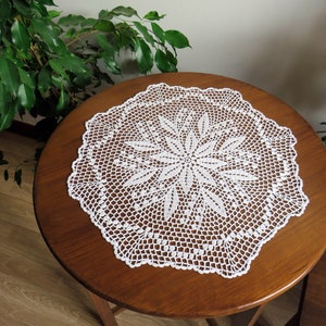 Può includere: Un centrino bianco all'uncinetto fatto a mano con un disegno floreale, appoggiato su un tavolo rotondo di legno. Il centrino presenta un motivo floreale centrale circondato da un'intricata lavorazione traforata. Il tavolo è marrone scuro.