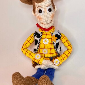 May include: Crocheted toy of Woody from Toy Story. He is wearing a yellow and red plaid shirt, a brown vest with a black and white pattern, a brown cowboy hat, and blue jeans. He has brown boots and a friendly smile.