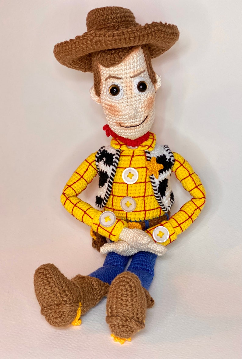 May include: Crocheted toy of Woody from Toy Story. He is wearing a yellow and red plaid shirt, a brown vest with a black and white pattern, a brown cowboy hat, and blue jeans. He has brown boots and a friendly smile.