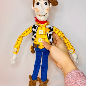 May include: A crocheted toy figure of Woody from Toy Story. He is wearing a brown cowboy hat, a yellow and red plaid shirt, a black and white vest, blue jeans, and brown cowboy boots.