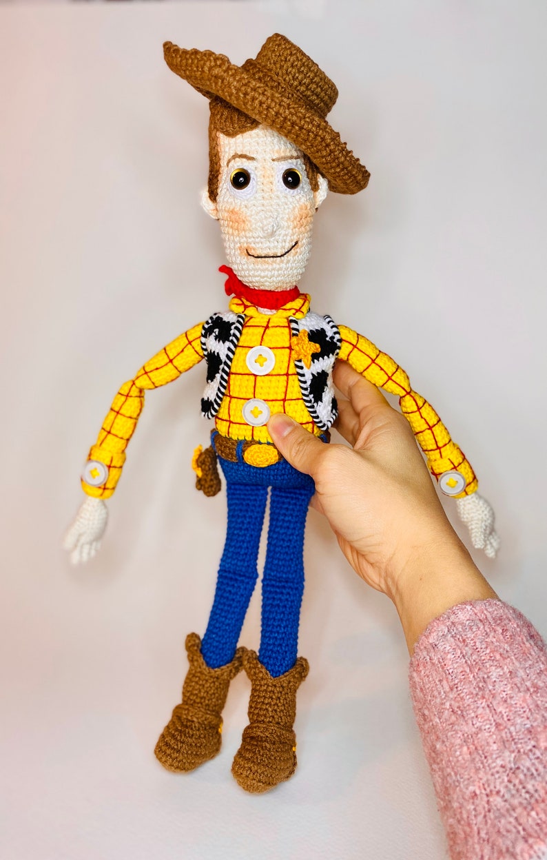 May include: A crocheted toy figure of Woody from Toy Story. He is wearing a brown cowboy hat, a yellow and red plaid shirt, a black and white vest, blue jeans, and brown cowboy boots.