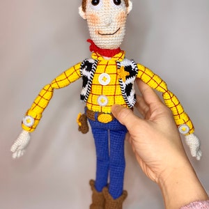 May include: Crocheted toy of Woody from Toy Story. He is wearing a yellow and red plaid shirt, a brown vest with a white star, blue jeans, and brown boots. He has brown hair and a friendly face.