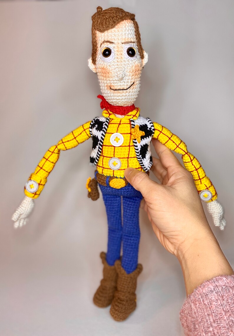 May include: Crocheted toy of Woody from Toy Story. He is wearing a yellow and red plaid shirt, a brown vest with a white star, blue jeans, and brown boots. He has brown hair and a friendly face.