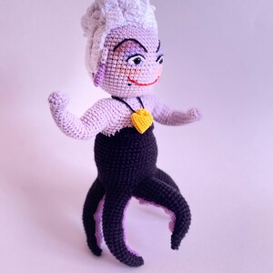 May include: A crocheted Ursula doll from The Little Mermaid. She has purple skin, white hair, and a black octopus tail. She is wearing a black dress and a yellow heart necklace.