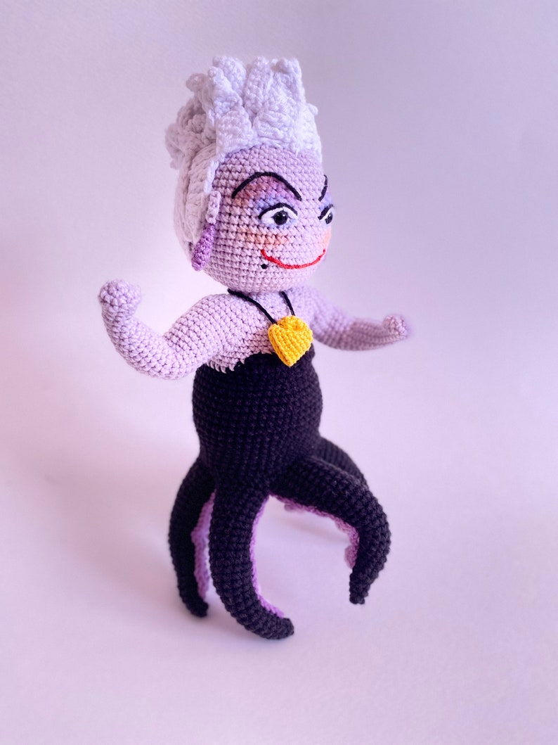 May include: A crocheted Ursula doll from The Little Mermaid. She has purple skin, white hair, and a black octopus tail. She is wearing a black dress and a yellow heart necklace.
