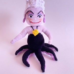 May include: A crocheted Ursula doll from The Little Mermaid. She has purple skin, white hair, and a black octopus tail. She is wearing a black dress and a yellow heart necklace.