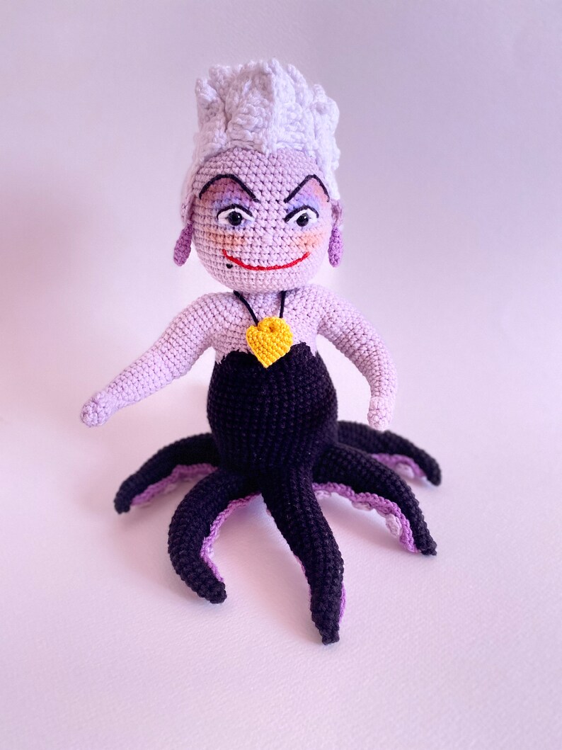 May include: A crocheted Ursula doll from The Little Mermaid. She has purple skin, white hair, and a black octopus tail. She is wearing a black dress and a yellow heart necklace.