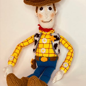 May include: A crocheted toy of Woody from Toy Story. He is wearing a brown cowboy hat, a yellow and red plaid shirt, a black and white vest, blue jeans, and brown boots. He has a red bandana around his neck and a sheriff's star on his vest.