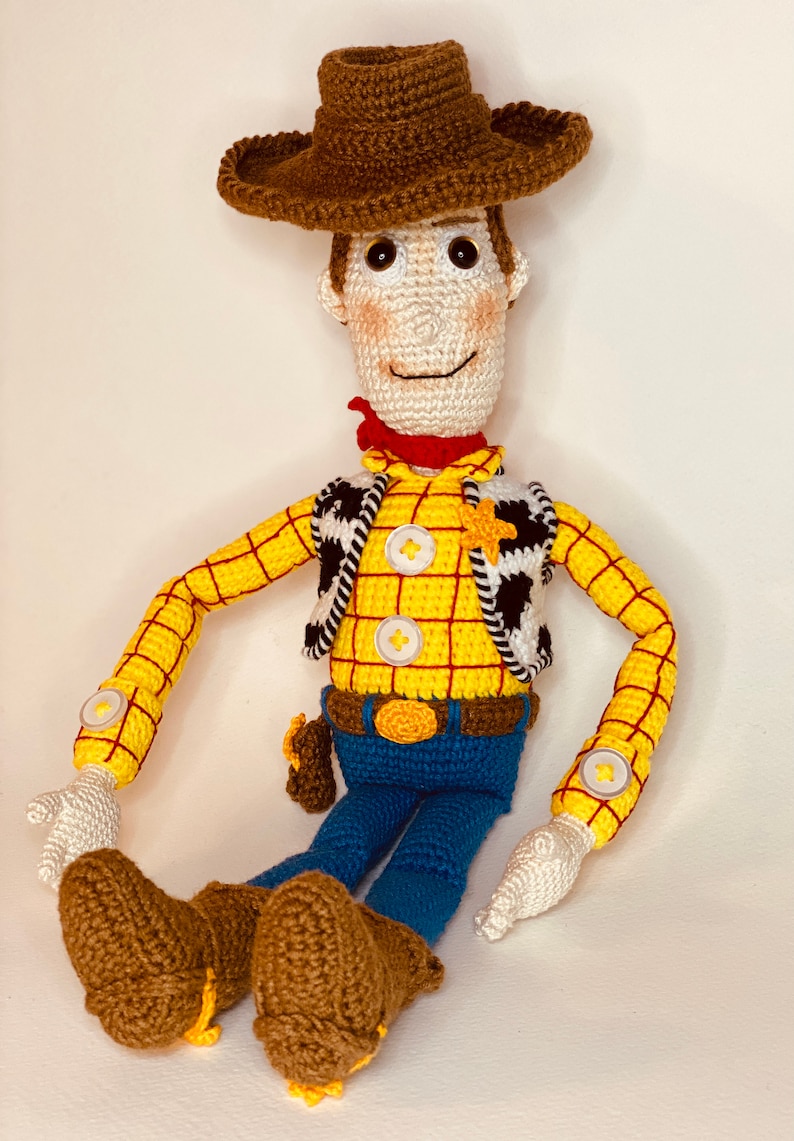 May include: A crocheted toy of Woody from Toy Story. He is wearing a brown cowboy hat, a yellow and red plaid shirt, a black and white vest, blue jeans, and brown boots. He has a red bandana around his neck and a sheriff's star on his vest.