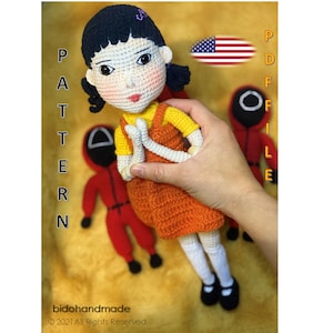 Peut inclure: Fichier PDF de modèle de crochet pour une poupée avec des cheveux noirs, une robe jaune et orange et des collants blancs. La poupée tient ses mains jointes en position de prière. Le modèle comprend un drapeau américain et les mots "PDF FILE" en texte noir.