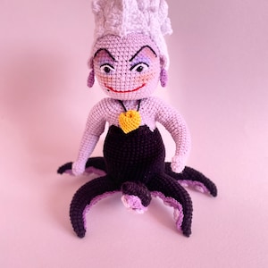 May include: A crocheted Ursula doll from The Little Mermaid. She has purple skin, white hair, and a black octopus tail. She is wearing a purple necklace with a yellow heart pendant.