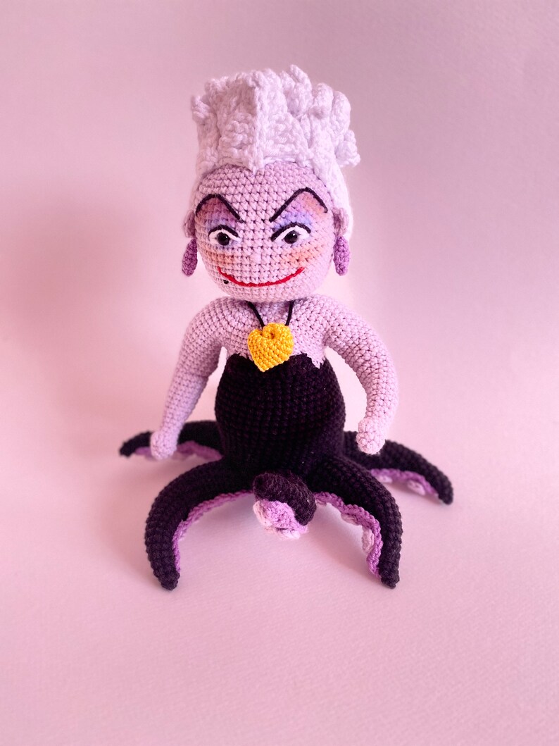 May include: A crocheted Ursula doll from The Little Mermaid. She has purple skin, white hair, and a black octopus tail. She is wearing a purple necklace with a yellow heart pendant.