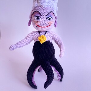 May include: A crocheted Ursula doll with purple and black yarn. She has a white crocheted crown, a yellow heart necklace, and a mischievous expression.