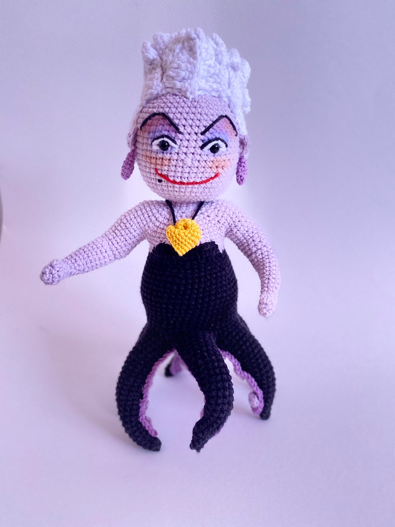 May include: A crocheted Ursula doll with purple and black yarn. She has a white crocheted crown, a yellow heart necklace, and a mischievous expression.