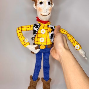 May include: A crocheted toy of Woody from the Toy Story franchise. Woody is wearing a yellow and red plaid shirt, a brown vest with a black and white pattern, a brown cowboy hat, blue jeans, and brown cowboy boots.