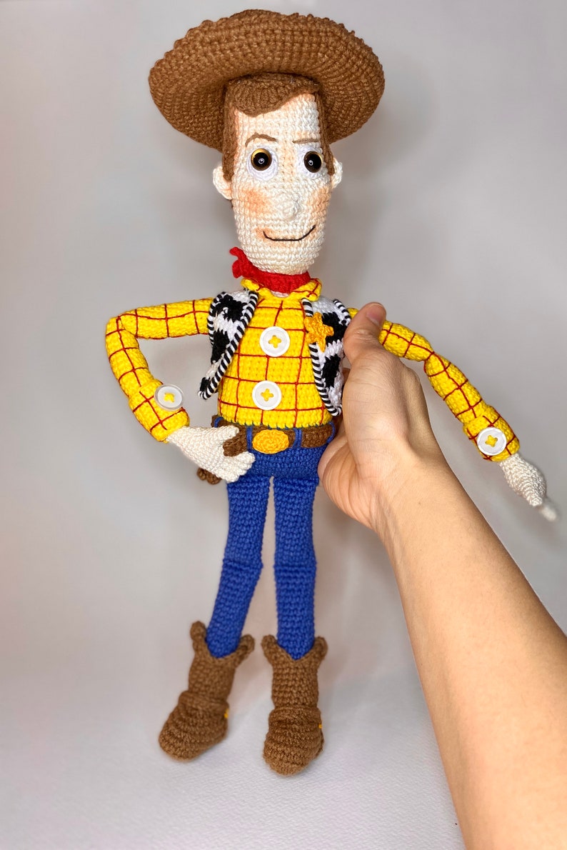 May include: A crocheted toy of Woody from the Toy Story franchise. Woody is wearing a yellow and red plaid shirt, a brown vest with a black and white pattern, a brown cowboy hat, blue jeans, and brown cowboy boots.