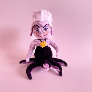 May include: A crocheted Ursula doll from The Little Mermaid. She has purple skin, a white crocheted crown, and a yellow heart necklace. She is sitting with her octopus tentacles spread out.