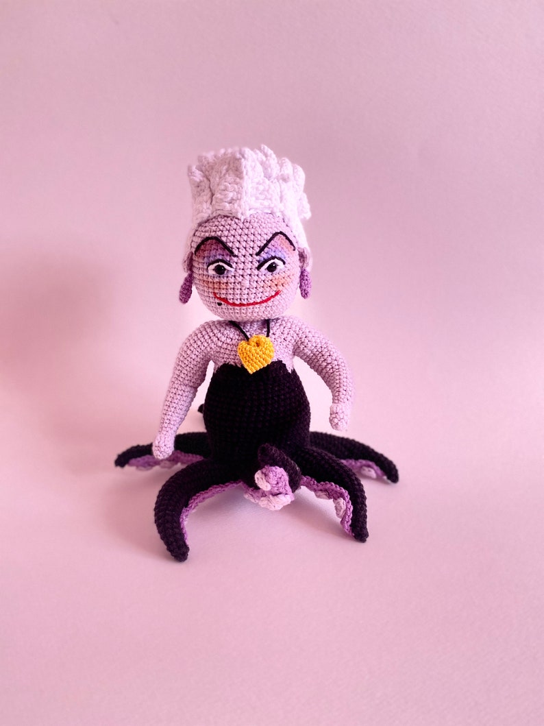 May include: A crocheted Ursula doll from The Little Mermaid. She has purple skin, a white crocheted crown, and a yellow heart necklace. She is sitting with her octopus tentacles spread out.